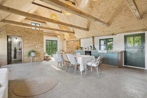 Een ruime kamer met een hoge plafond, houten balken, een eethoek met een tafel en stoelen, een keuken met een spoelbak en kastjes, en een groot raam.