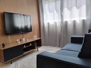 A modern living room with a flat-screen TV mounted on a wooden panel wall, a dark-colored sofa, and a small white vase on a wooden console table. - Fully furnished apartment near the south beach of João Pessoa. (João Pessoa)