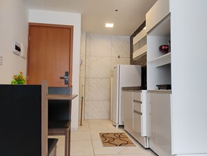 A compact kitchen with a wooden door, white cabinets, a white refrigerator, and a small dining table. - Fully furnished apartment near the south beach of João Pessoa. (João Pessoa)