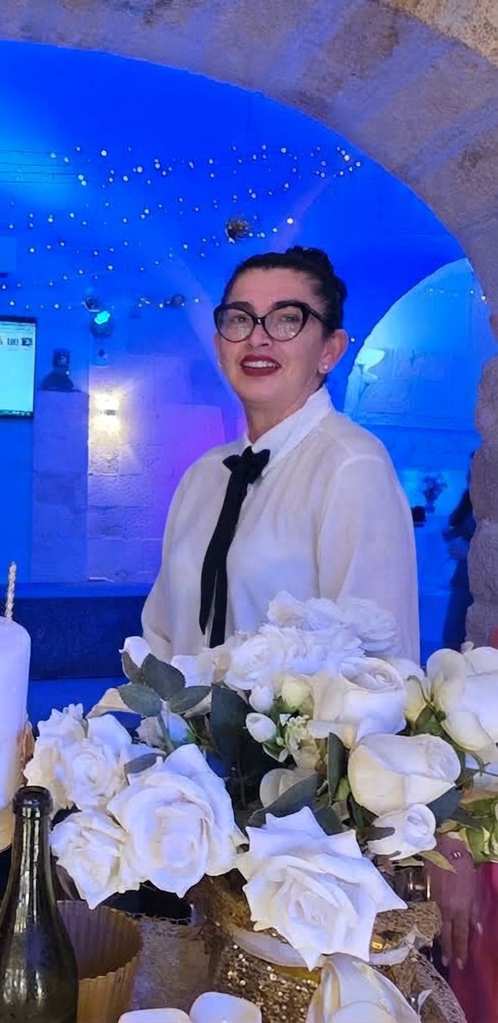 A person standing in front of a table with white flowers and a bottle, in a room with blue lighting and a stone wall.