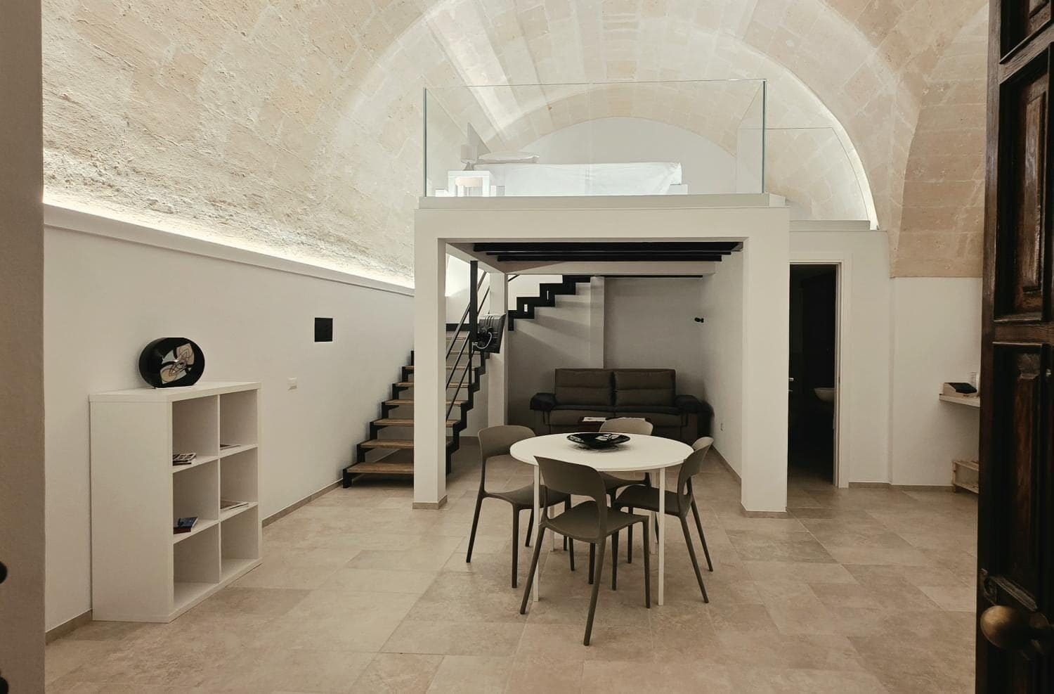 A modern interior with a staircase leading to a loft, a dining area with chairs, and a bookshelf.