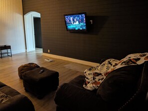 En stue med et tv monteret på en mørk væg, en brun sofa og en lille skammel med en fjernbetjening.