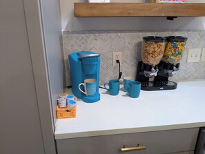 En køkkenbordplade med kaffemaskine, to kopper, en cornflakesdispenser og en frugtbeholder.