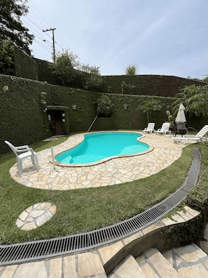 Piscina com deck de pedra, cercada por gramado e um arbusto.