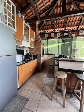 A kitchen with wooden cabinets, a refrigerator, a microwave, and a window overlooking greenery. - House of the Master Brewer (Blumenau)