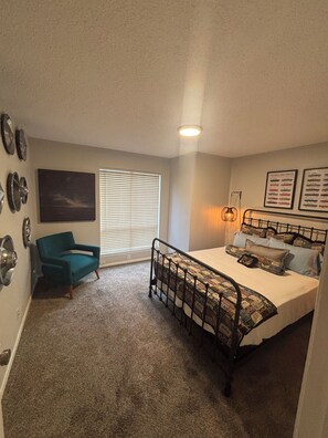 A bedroom with a bed, a chair, a window with blinds, and wall decorations. -     Cozy 1BR Near Dallas Medical District  - Furnished + Utilities (Dallas)