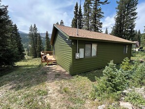 Eine kleine Holzhütte mit einer Holzterrasse und einem Fenster, umgeben von Bäumen und einem Berg im Hintergrund.
