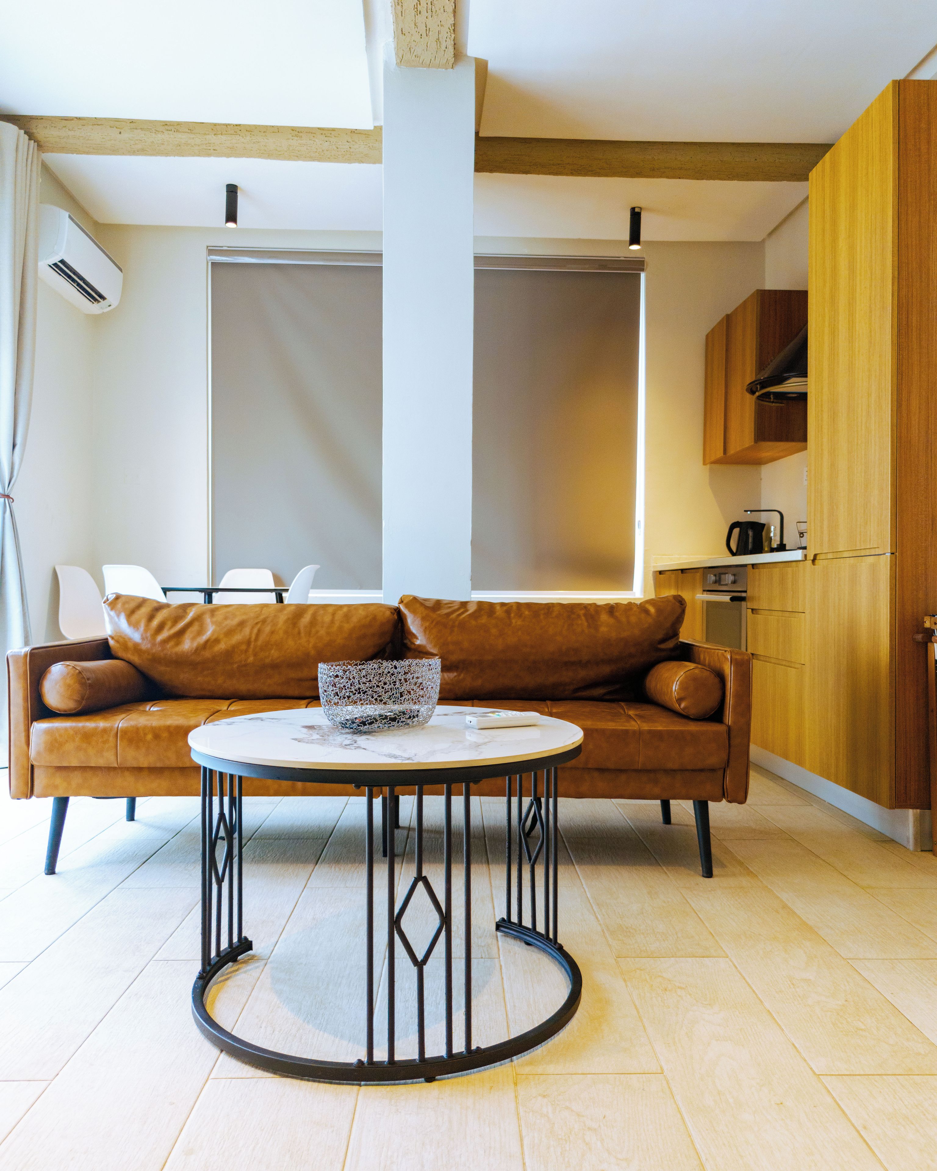 Uma sala de estar moderna com um sofá de couro marrom, uma mesa de centro redonda e uma área de cozinha com armários de madeira.