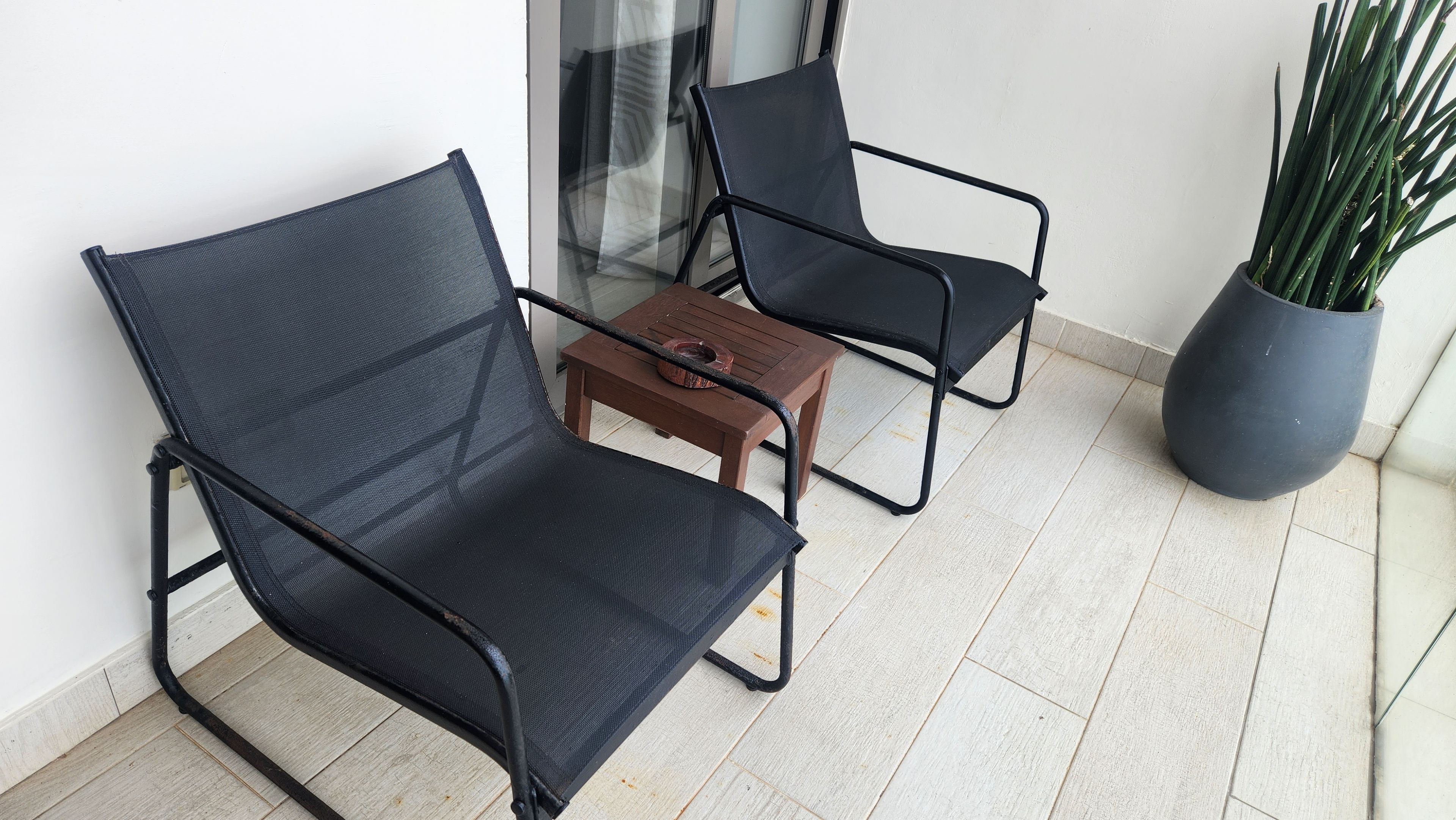 Two black mesh chairs with metal frames, a small wooden table with a bowl, and a potted plant on a balcony with light-colored flooring.