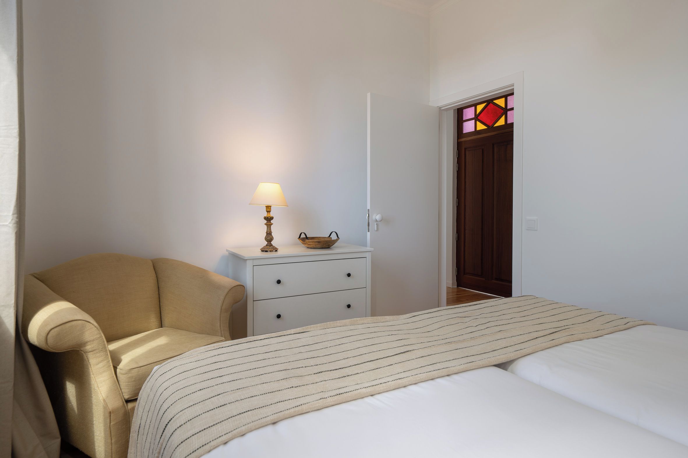 Une chambre à coucher avec un lit, une chaise, une table de chevet avec une lampe et une porte ornée d’un vitrail.