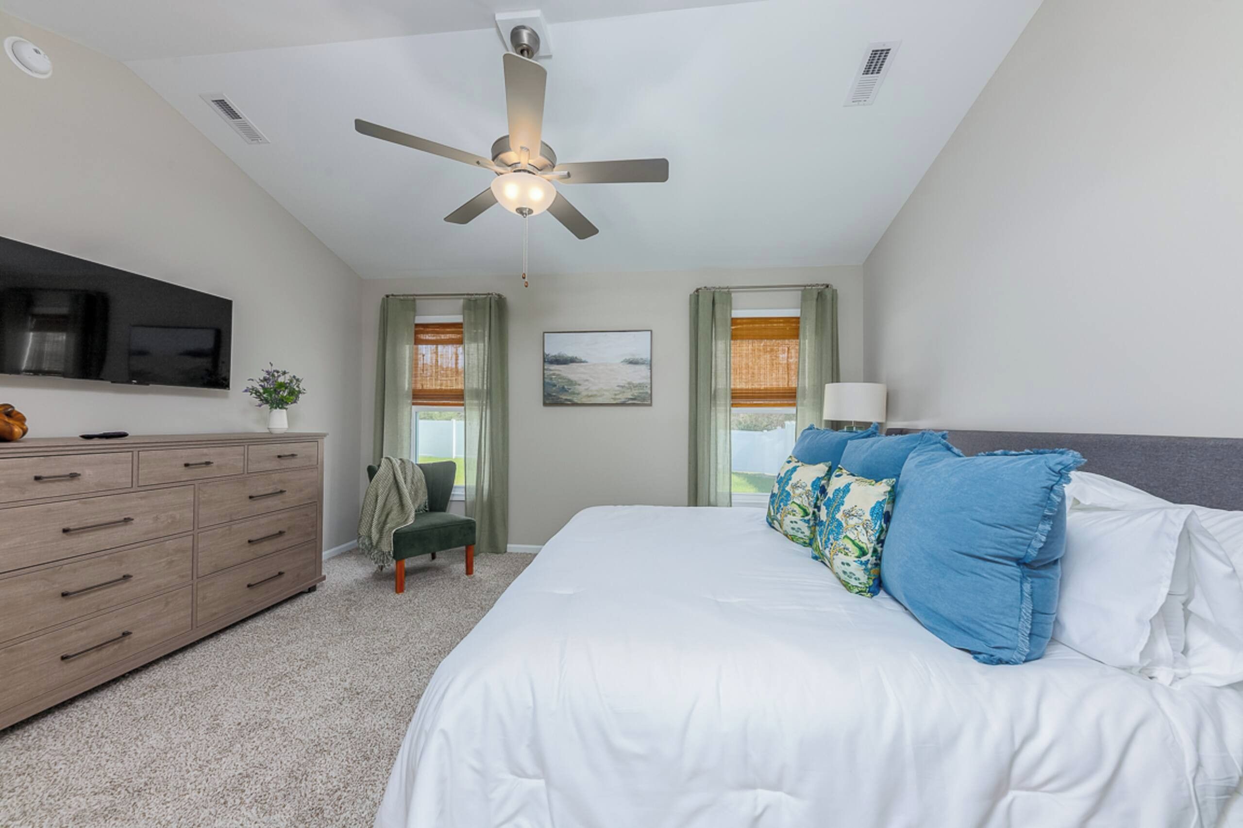 A bedroom with a bed, a dresser, a ceiling fan, a television, and windows with curtains.