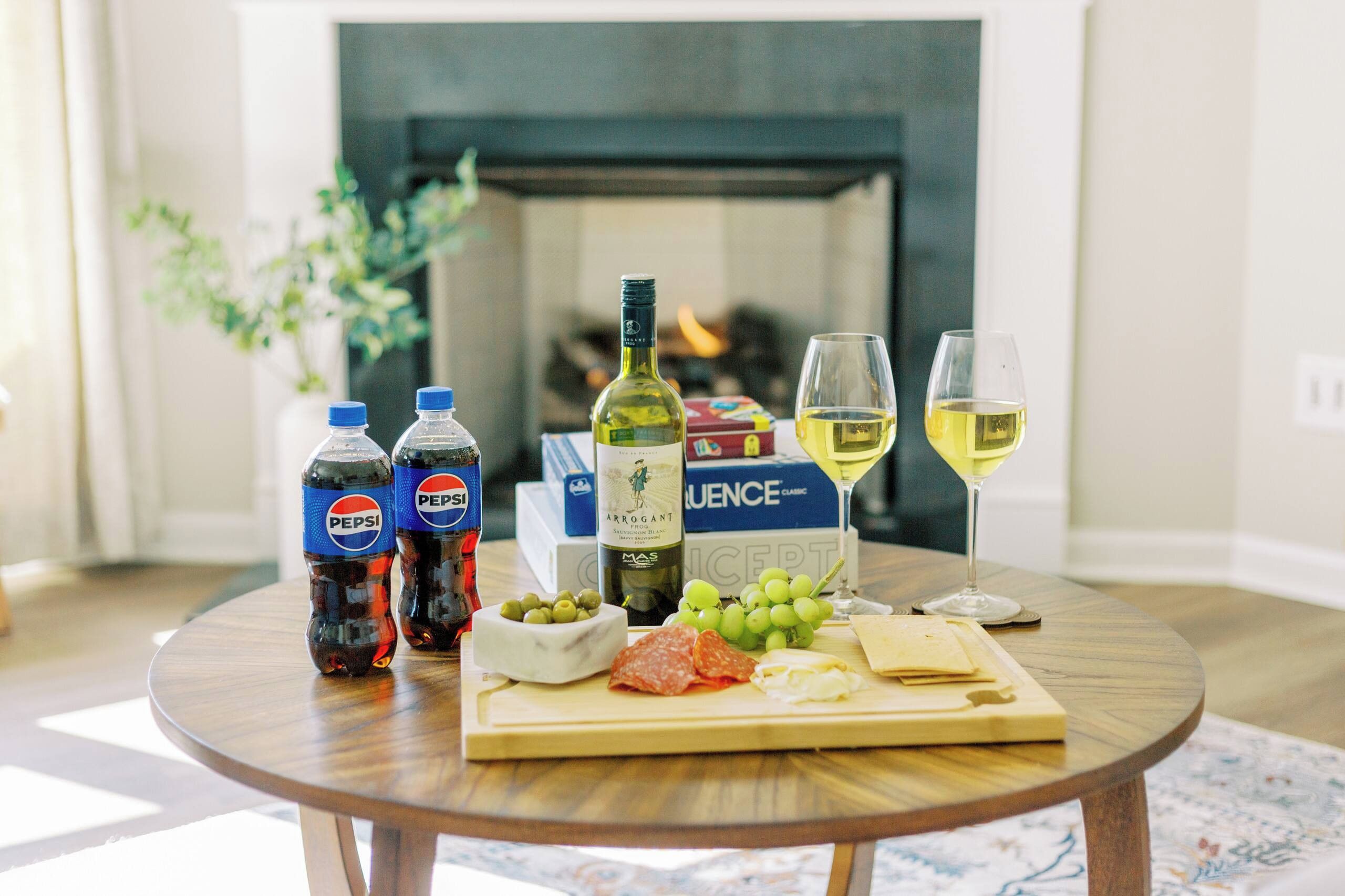 A round wooden table with a bottle of Arrogant wine, two bottles of Pepsi, a glass of white wine, a bowl of grapes, and a cheese board with various cheeses and meats.