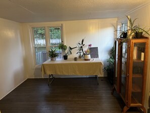 A room with a table covered by a tablecloth, flanked by windows with white blinds, a wooden cabinet with glass doors, and several potted plants.