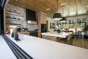 A modern kitchen with a large island, wooden ceiling, stone wall backsplash, and a dining area with natural light. - NEW ! Bear Lake Lodge - Private pond/Hidden speakeasy/pool/pedal boat/kayaks (Broken Bow)