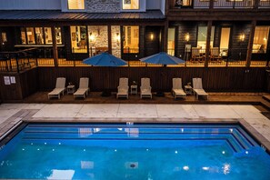 Ein Schwimmbecken mit Liegestühlen und Sonnenschirmen, umgeben von einer Holzterrasse und einem Gebäude mit Fenstern und Lichtern.
