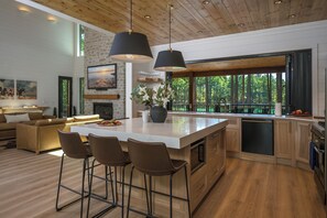 Eine moderne Küche mit einer großen Kochinsel, Barhocker, einer Essecke mit einer Couch und Blick auf den Wald durch große Fenster.