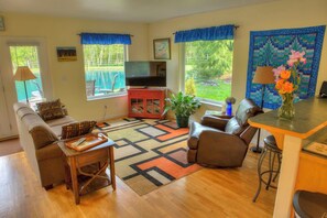 A living room with a sofa, armchair, coffee table, TV stand, and a dining area with a table and chairs. - Come Relax at Mountain Springs Cabin w/ Your Own Private Pond (Mount Vernon)