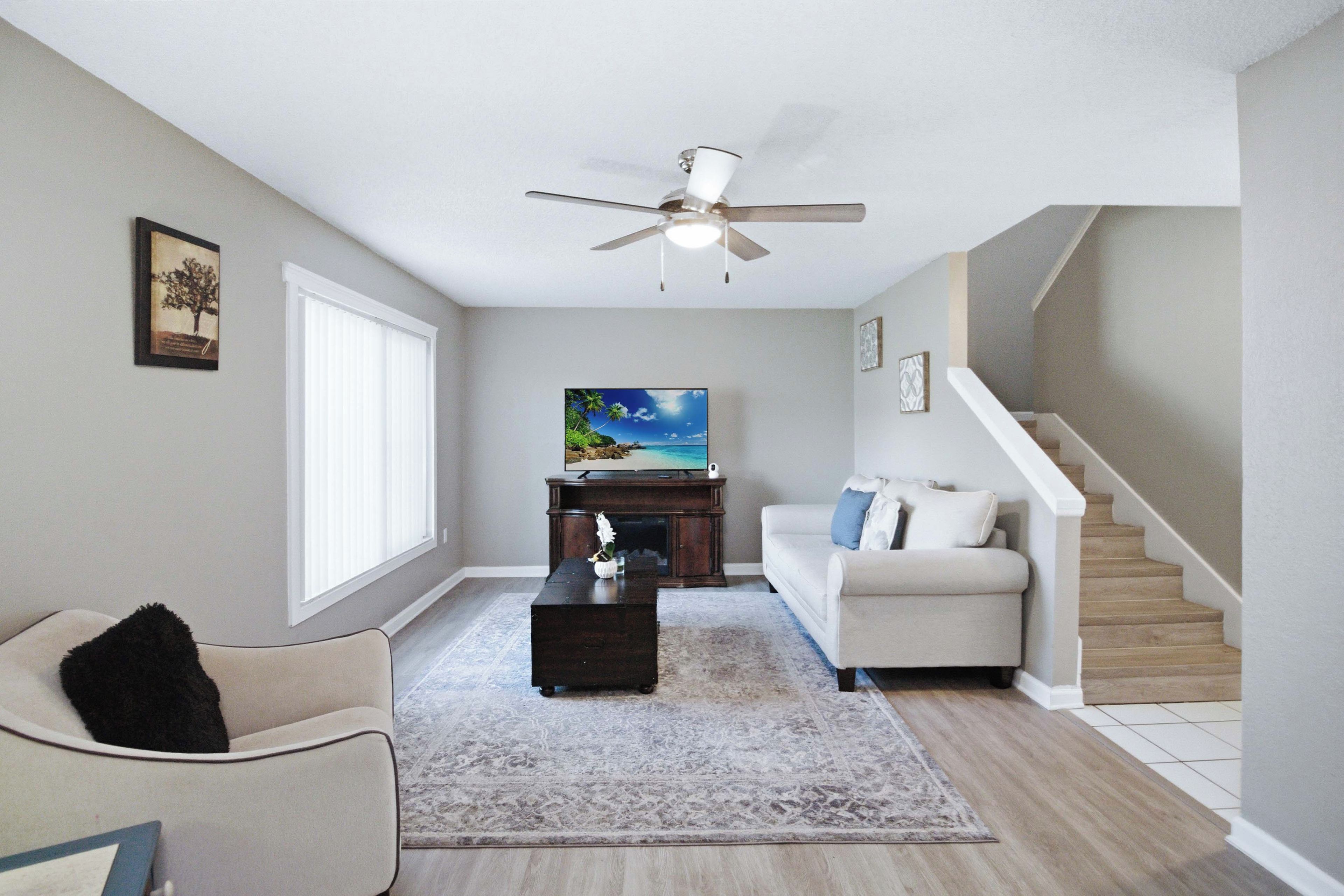 A living room with a sofa, a coffee table, a TV on a stand, a ceiling fan, and a staircase.