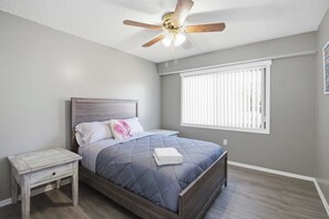 Une chambre à coucher comprenant un lit, une table de chevet, un ventilateur de plafond, une fenêtre avec stores et un interrupteur fixé au mur.