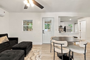 A modern living room with a black sofa, a round dining table with chairs, a ceiling fan, and a view of the kitchen through an open door. - 5BR , Sleeps 14 , Central to Downtown & Tybee (Savannah)