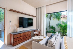 A modern living room with a flat-screen TV mounted on the wall, a wooden console table, a sofa with cushions, and a view of palm trees through large windows. - Beachfront 5BR Villa w/ Pool, big garden, Ocean Views & Premier Golf Membership (Punta de Mita)