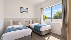 A hotel room with two beds, a nightstand, a lamp, a framed picture on the wall, and a large window with curtains. - Fingal Tide House - Spacious 3BR Beachside Getaway (Fingal Bay)