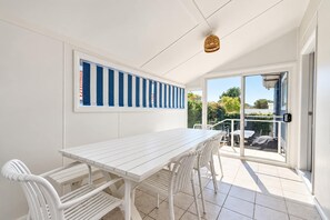 Une table à manger blanche avec des chaises, une fenêtre avec des stores à rayures bleues et blanches, et une vue sur la verdure à l’extérieur.