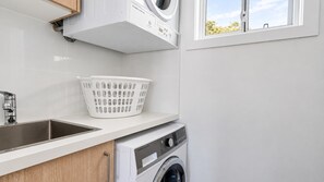 A laundry area with a washing machine, a sink, and a white laundry basket. - Fingal Tide House - Spacious 3BR Beachside Getaway (Fingal Bay)