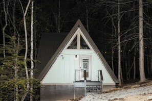 A small, triangular cabin with a door and a small porch, surrounded by trees. - Twilight at Lux in the Gorge - Hot Tub - Fishing  - Shared Pond with Kayaking, Swimming, & Pedal Boat - Game Room with Pool Table, Foosball, Ping Pong, Air Hockey, and More! (Campton)