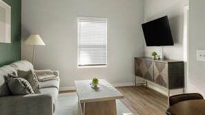 A living room with a sofa, a coffee table, a TV on a stand, and a window with blinds. - Landing Stylish 1bd, Clubhouse, Gym (Raleigh)