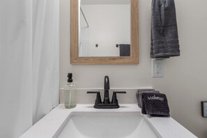 Une salle de bain avec un lavabo blanc, un miroir encadré en bois, un robinet noir, un distributeur de savon et un porte-serviettes avec une serviette foncée.