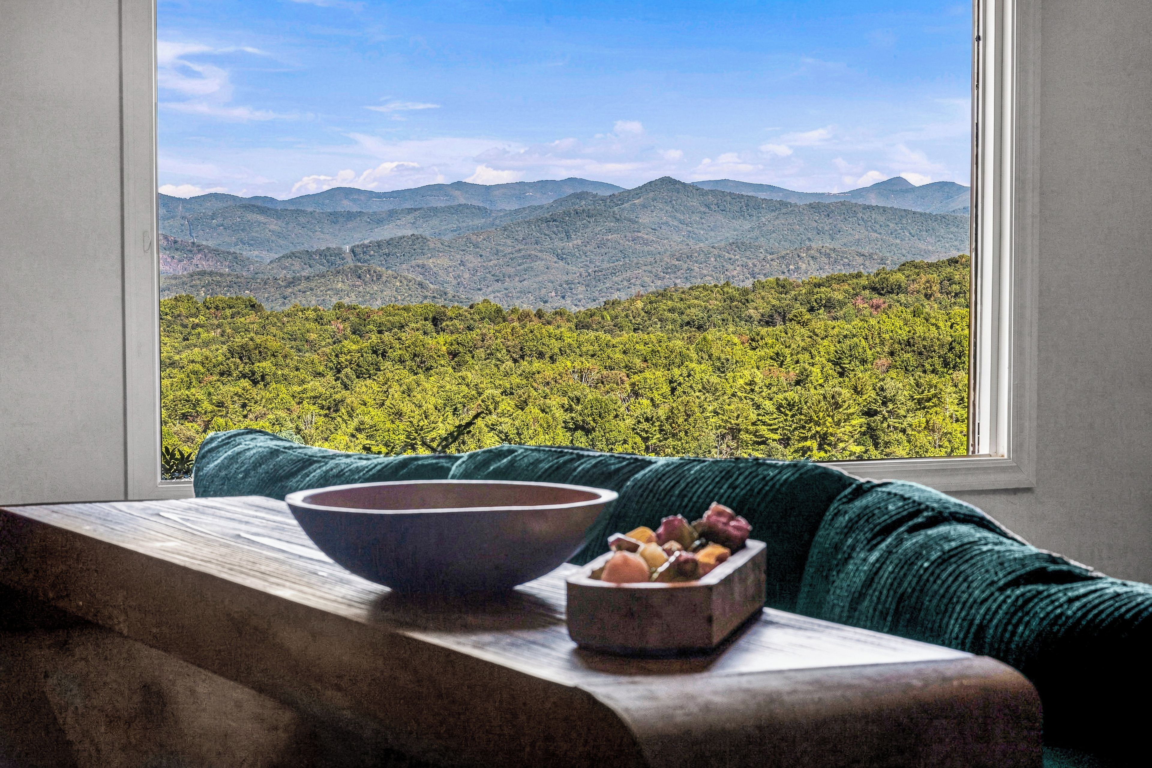 A window with a view of a mountainous landscape and a table with a bowl and a fruit basket.