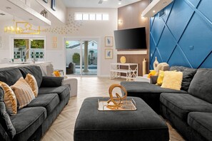 A modern living room with dark grey sofas, a coffee table with a decorative metal piece, and a large flat-screen TV mounted on a blue wall. - FreePoolHeat|HotTub|Sleeps18|Arcade|ThemeRooms (Mesa)