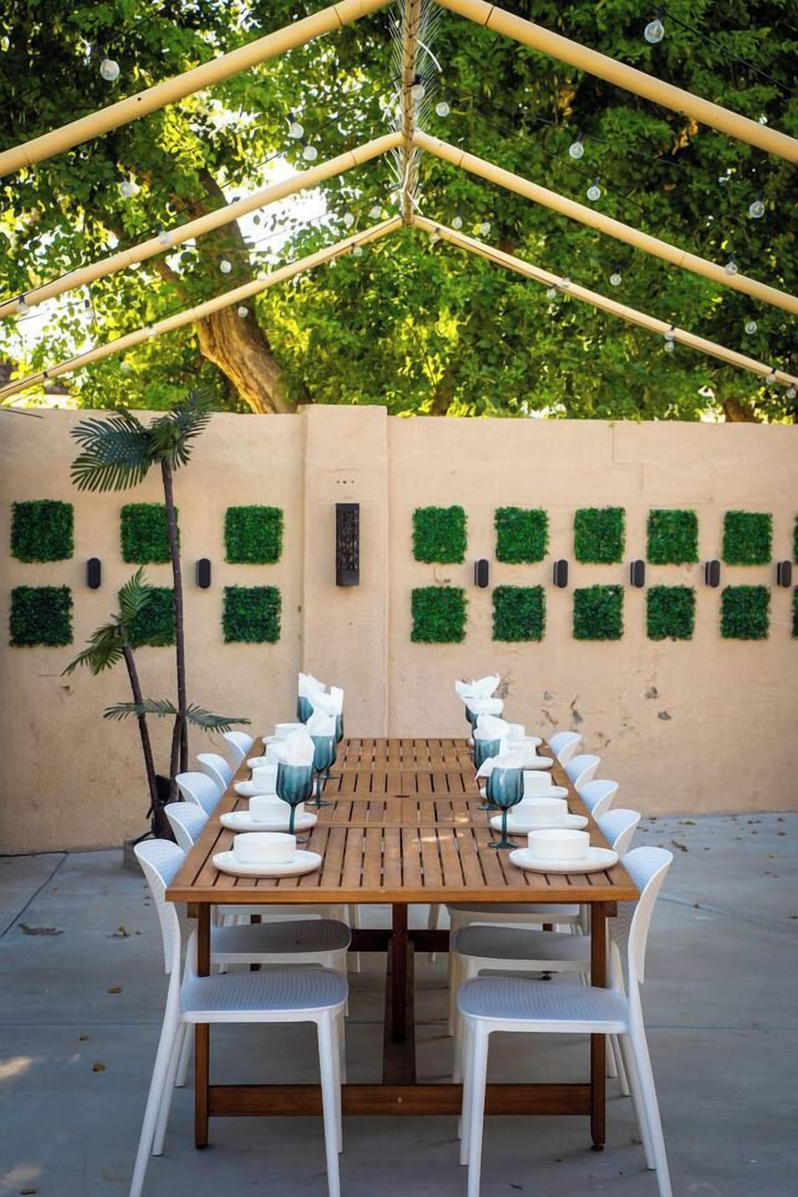 Un espace de restauration en plein air avec une longue table en bois et des chaises blanches, entouré d’un mur beige agrémenté de décorations végétales vertes.