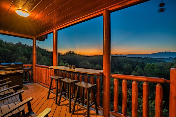 A wooden deck with a barbecue grill, bar stools, and a view of a forested landscape. - Mountain Views, 2 King Suites, Hot Tub, Cabin with AC in peaceful Sevierville (Sevierville)
