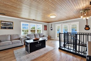 Ein geräumiges Wohnzimmer mit Holzboden, einem großen Fenster mit Jalousien, einem grauen Sofa, einem schwarzen Stauraum und einer Treppe mit schwarzer Geländer.