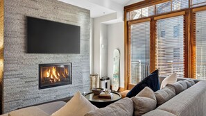 A living room with a fireplace, a flat-screen TV mounted on a textured wall, and a large window with blinds. - The Iron Horse by Avantstay Mountain Views Pool & Spa Walk to Gondola Skating Rink & Shops (Truckee)