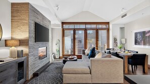 A modern living room with a fireplace, a television, and a dining area with a table set for two. - The Iron Horse by Avantstay Mountain Views Pool & Spa Walk to Gondola Skating Rink & Shops (Truckee)