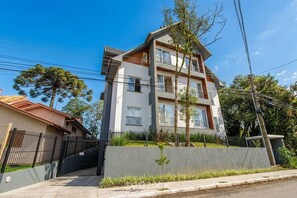 A multi-story residential building with a gated entrance, a small garden area, and a clear sky. - Apt. w/ Fireplace, City View & Garage Cun0102 (Gramado)