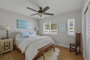 Un dormitorio con una cama, una mesita de noche, una silla, un ventilador de techo y ventanas.