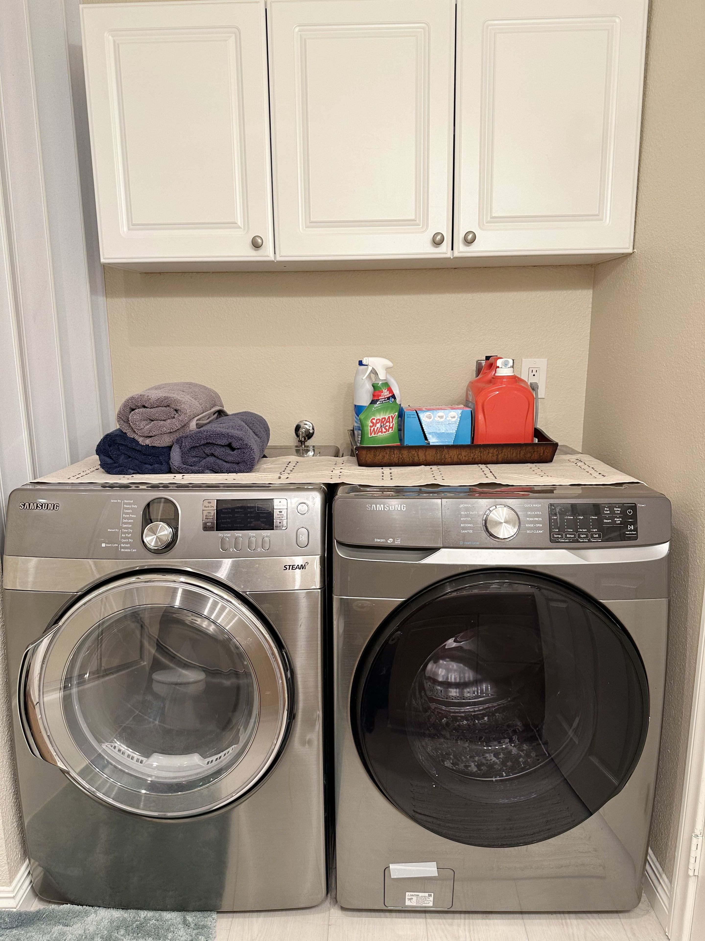 A laundry area with two front-loading washers, a drying rack with folded towels, and a shelf with cleaning supplies.