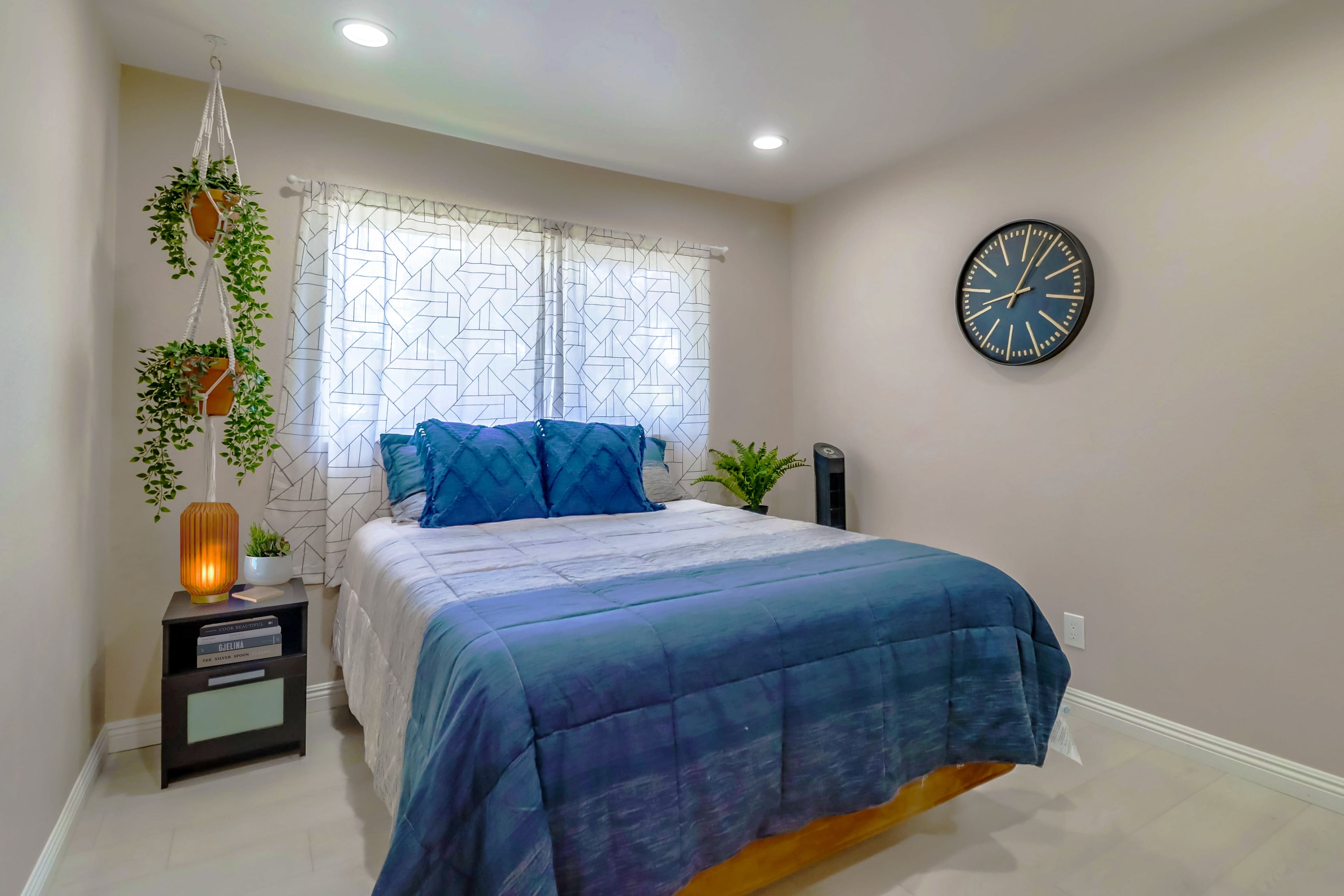 A bedroom with a bed, a wall clock, a bedside table with a lamp, and a hanging plant.