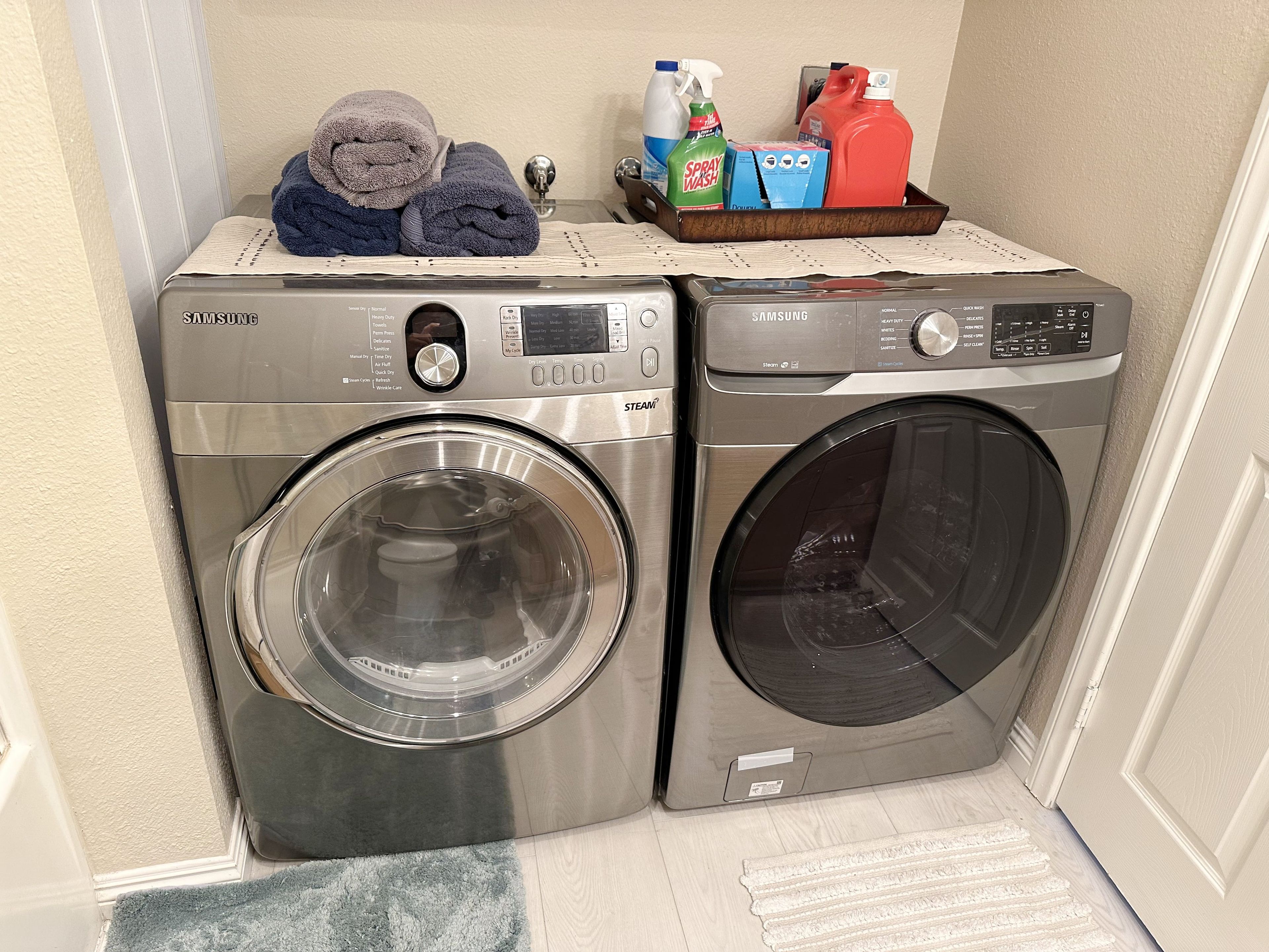 A laundry area with two front-loading washing machines, a rack with folded towels, and cleaning supplies.