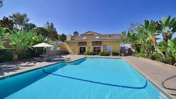 Una piscina rectangular con superficie de agua azul cristalina, rodeada por una solana empedrada. Hay sillas de descanso y una pequeña zona de estar cubierta. La piscina está adyacente a un edificio con fachada amarilla y techo plano.
