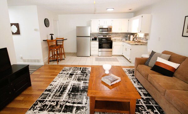 A modern living room with a wooden coffee table, a brown sofa with patterned cushions, a kitchen area with white cabinets and stainless steel appliances, and a dining area with wooden chairs.