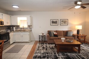 A modern kitchen with white cabinets, a microwave, and a sink. There is a living area with a brown sofa, a coffee table, and a rug. Two framed pictures are on the wall.