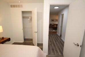 A hotel room with a bed, a nightstand, a lamp, and a door leading to a hallway with a wooden floor and a small kitchen area.