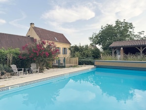 Ein Schwimmbad mit gefliestem Deck, ein Haus mit rotem Dach und ein Pavillon.