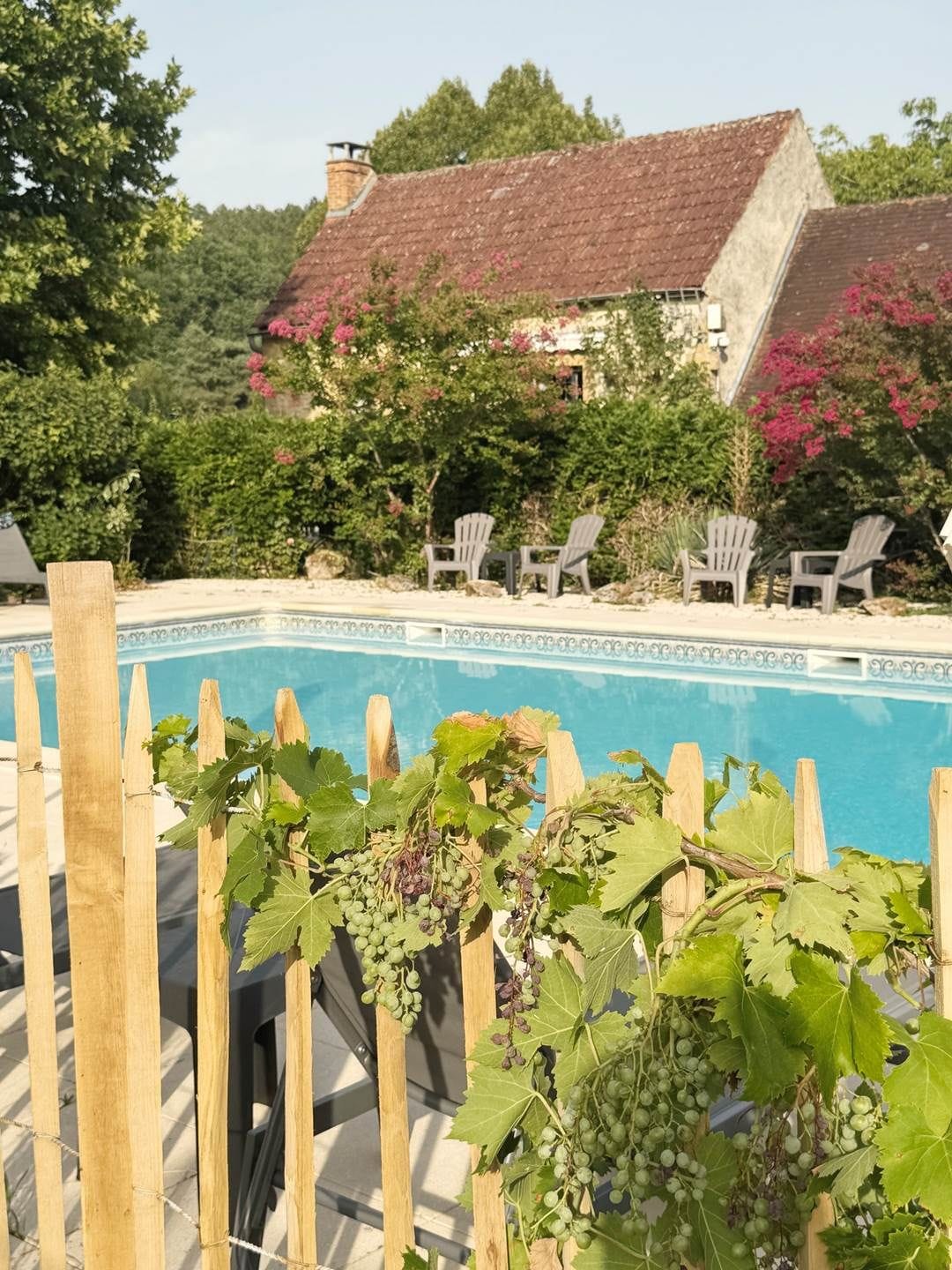 Zona de piscina amb tanca de fusta, filera de cadira i un edifici amb teulada vermella al fons.
