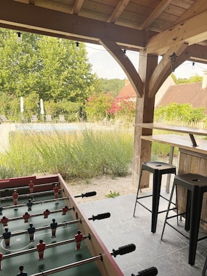 A foosball table under a wooden structure with a view of a garden and a pool. - La Violette – Spacious holiday home with private terrace and views (Meyrals)